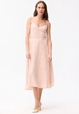 Donna che indossa un vestito midi rosa chiaro senza maniche con spalline sottili e scarpe slingback beige con tacco alto, in piedi davanti a uno sfondo bianco.