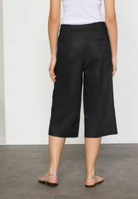 Pantalones culotte de lino negros con corte suelto, de longitud hasta el tobillo y un solo detalle de bolsillo trasero. Sandalias metálicas complementan el conjunto.