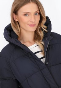 Jachetă puffer bleumarin cu glugă, textură matlasată, fermoar frontal și închidere cu capse. Asortată peste un tricou cu dungi.