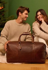 Brun läderväska med dubbla övre handtag, dragkedjestängning och yttre ficka; slät textur, rundad form och minimalistisk design.