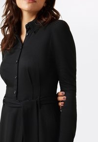 Vestito nero con bottoni, maniche lunghe, vita cinta, tessuto liscio e colletto morbido. Presenta tasche laterali discrete e dettagli con bottoni.