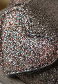 Zapato gris brillante con un destacado parche de purpurina en forma de corazón. La textura es suave y presenta detalles de purpurina multicolor cosidos a lo largo del borde.