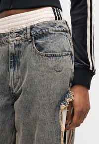 Άτομο που φοράει τζιν με acid-wash μπλε χρώμα και ξεφτισμένη πλαϊνή σχισμή, μαύρο αθλητικό μπουφάν Adidas και λευκή μέση με μαύρες ρίγες.