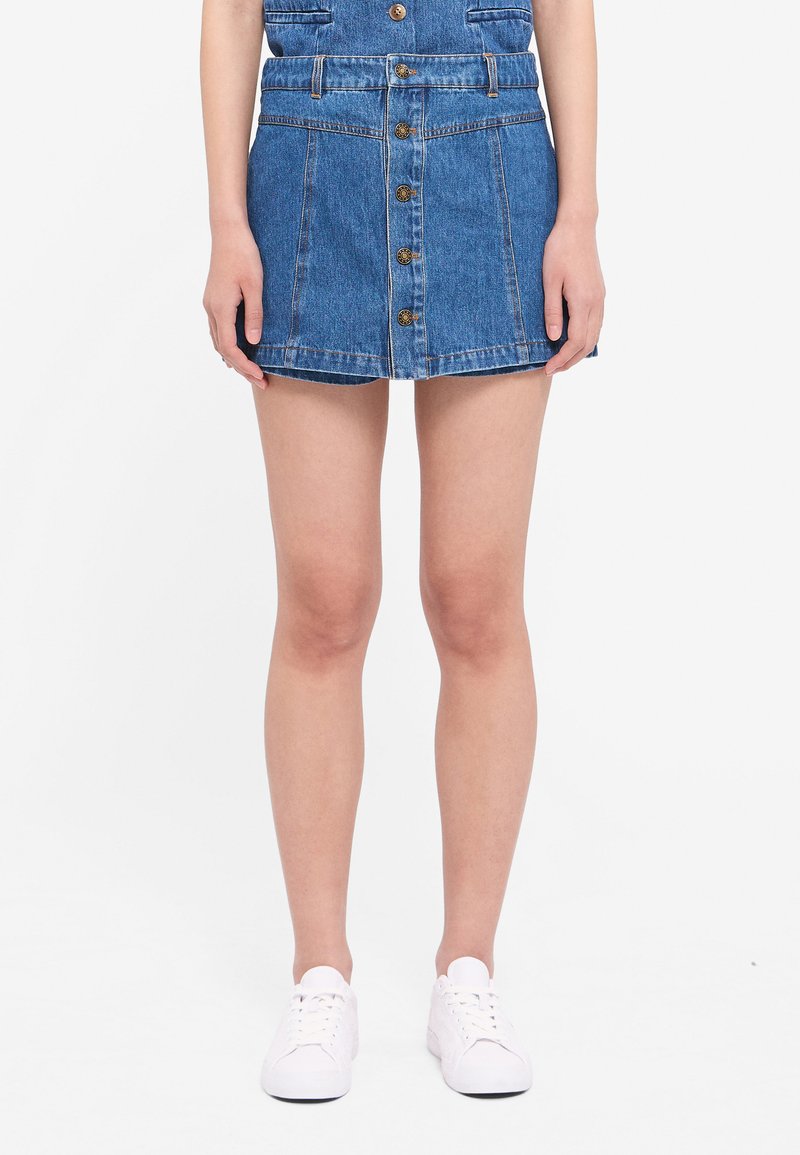 Denim skirt with a fitted waist, five front buttons, two front pockets, and a slightly flared hem. Worn with white sneakers.