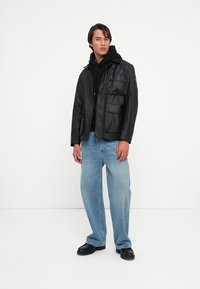 Black leather jacket with multiple front pockets, worn over a black hoodie. Light blue jeans and black shoes complete the look.