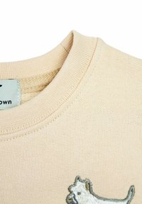 Beżowa bawełniana bluza z okrągłym dekoltem i prążkowanym wykończeniem. Posiada mały haftowany wzór psa w odcieniach szarości i bieli w dolnym lewym rogu.