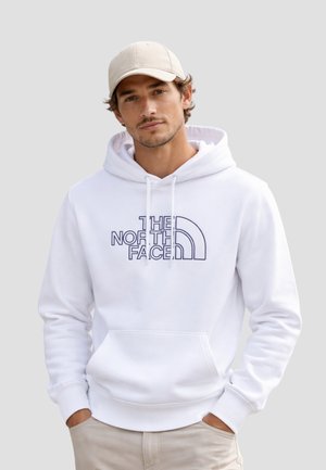 Uomo con felpa bianca con logo "The North Face", cappellino beige e pantaloni beige, in piedi con le mani nelle tasche.