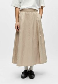 Personne portant une jupe midi beige taille haute boutonnée, des chaussettes blanches, des mocassins noirs et une chemise blanche à manches courtes, une main dans la poche.