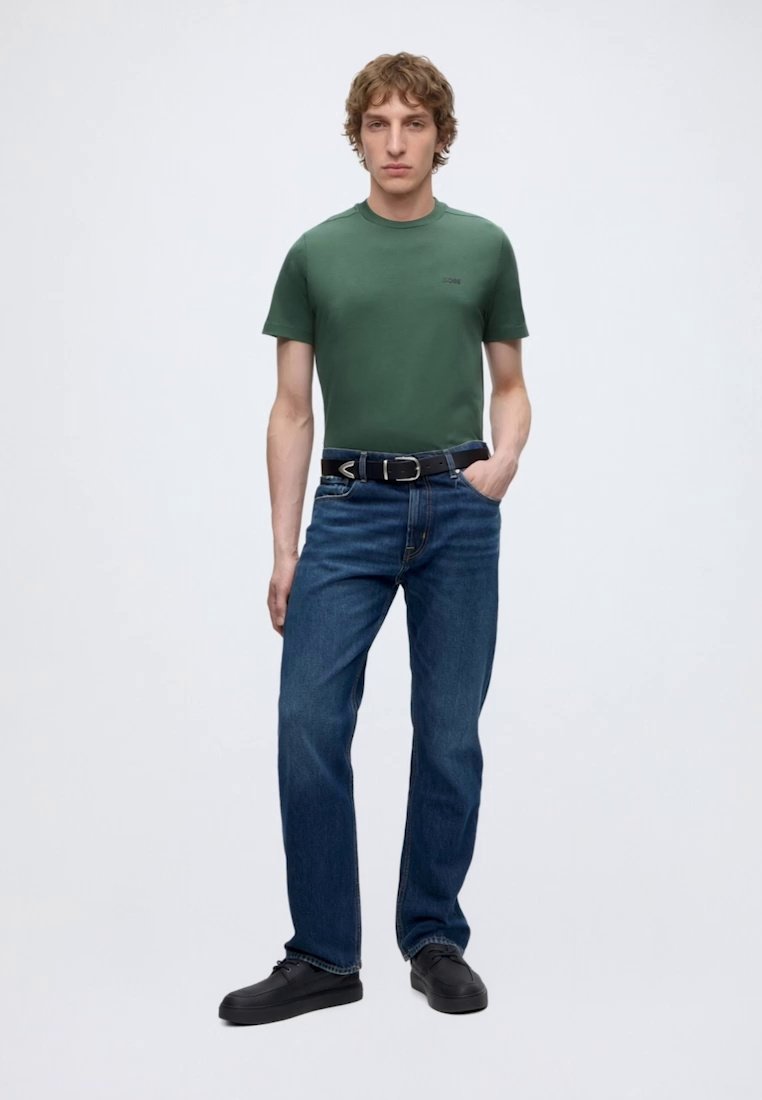 Jeune homme aux cheveux bouclés portant un t-shirt vert, un jean bleu, une ceinture noire et des chaussures noires, debout avec une main dans la poche.