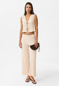 Beige, strukturiertes Cropped-Top mit V-Ausschnitt und vorderem Bindeband, kombiniert mit passenden weit geschnittenen Hosen. Ergänzt mit einer kleinen braunen Clutch.