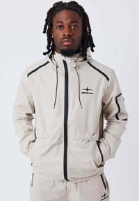 Giacca beige chiaro con dettagli neri, zip frontale, cappuccio e polsini elasticizzati. Presenta un logo sul petto e diverse tasche. Texture liscia.