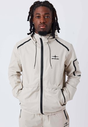 Veste beige clair avec des accents noirs, fermeture éclair devant, capuche et poignets élastiques. Présente un logo sur la poitrine et plusieurs poches. Texture lisse.