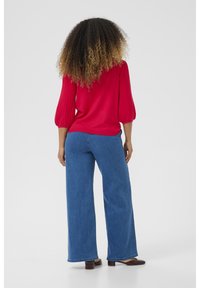 Rote, gerippte Bluse mit Puffärmeln, kombiniert mit weit geschnittenen blauen Jeans und braunen Schuhen, von hinten vor einem schlichten Hintergrund betrachtet.