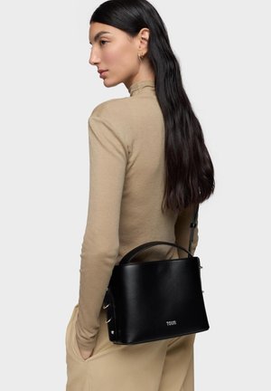 Donna con lunghi capelli scuri indossa un dolcevita beige e pantaloni, portando una borsa Tous nera con tracolla sulle spalle.