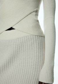 Gestreiftes cremefarbenes Strickkleid mit taillierter Silhouette, asymmetrischem Design und langen Ärmeln, das strukturierte Muster präsentiert.