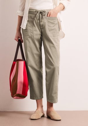 Persona che indossa pantaloni corti beige chiaro con tasche frontali e coulisse, scarpe slip-on beige, che tiene una borsa a righe rossa e rosa.