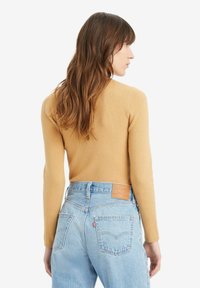 Sennepfarbener, gerippter Langarmoberteil, kombiniert mit hellblauen, hoch taillierten Jeans, die ein Lederpatch und zwei Gesäßtaschen aufweisen.