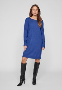 Stickad blå klänning med långa ärmar och ribbade manschetter, med en rundad halsringning. Bärs med svarta knähöga stiletto stövlar.