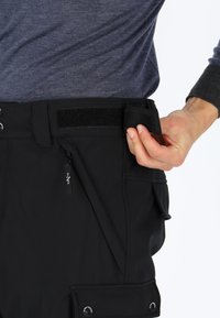 Zwarte broek met een gestructureerd oppervlak, met een klittenbandsluiting op de taille, een ritszak en drukknoopsluitingen aan de zijkanten.