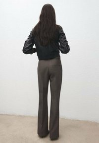 Kobieta o długich brązowych włosach stoi twarzą do białej ściany, ubrana w czarną skórzaną kurtkę, szare dopasowane spodnie i czarne szpilki z ostrym noskiem.