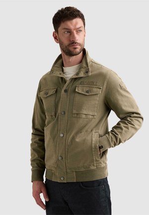 Homme aux cheveux courts et foncés portant une veste marron clair boutonnée avec des poches poitrine, debout, une main dans la poche, regardant à gauche.