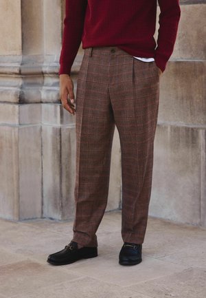 Brown plaid trousers made of textured fabric, featuring a loose fit and front pleats. Paired with black loafers.
