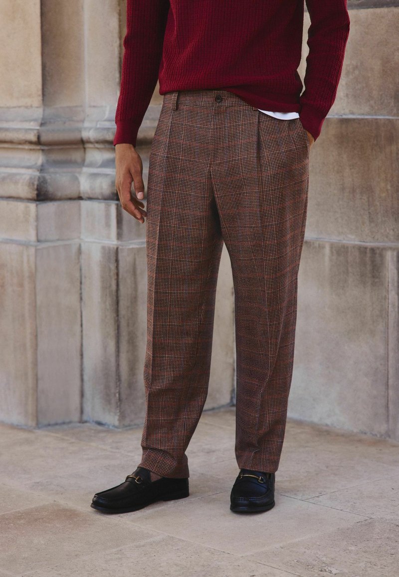 Brown plaid trousers made of textured fabric, featuring a loose fit and front pleats. Paired with black loafers.