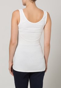 Nainen seisoo selkä kameraan päin, päällään valkoinen ilman hihoja oleva toppi ja tummat housut yksinkertaista taustaa vasten.