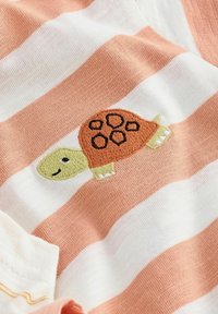 Blødt strikket stof med hvide og ferskenfarvede vandrette striber samt en lille broderet skildpadde med et orange skal og grøn krop.