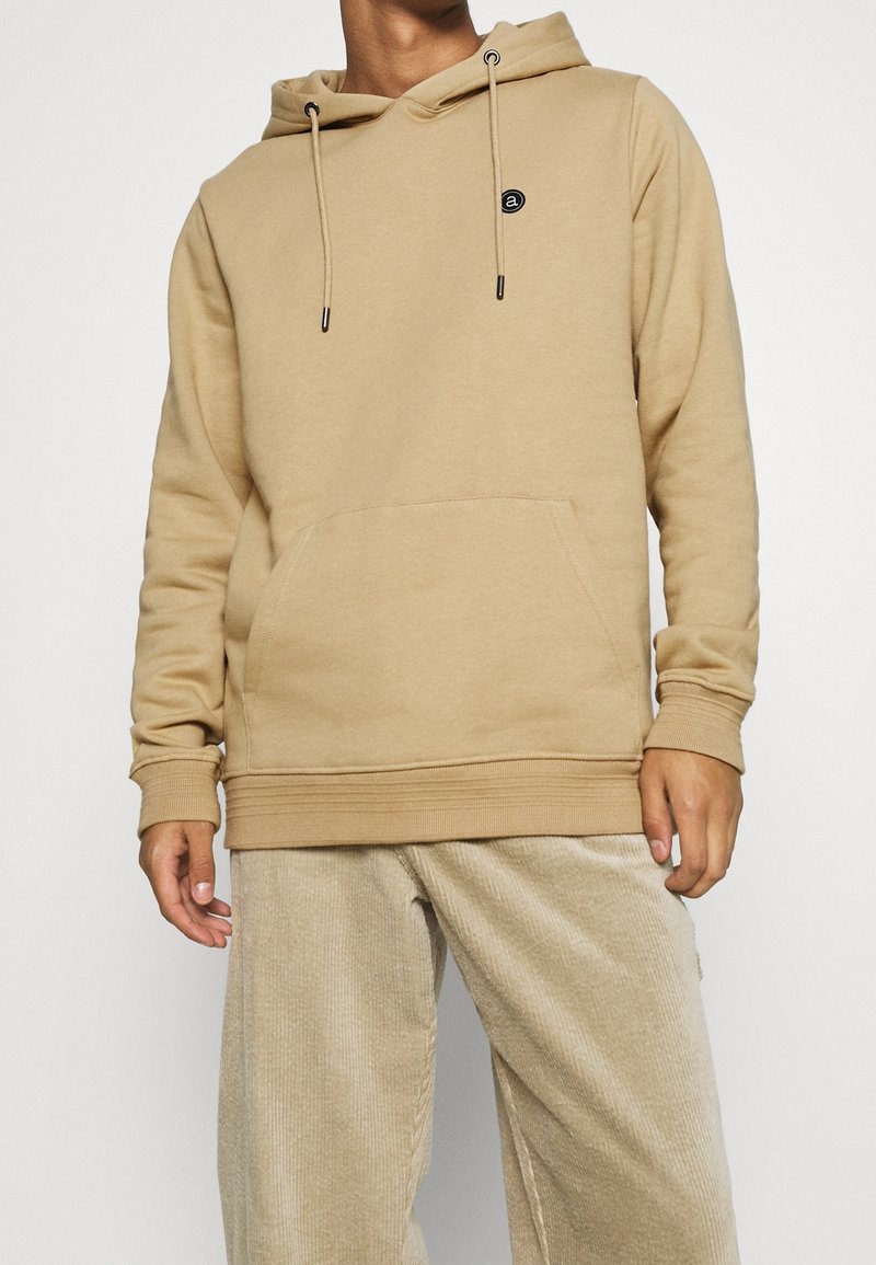 Felpa con cappuccio beige realizzata in morbido tessuto, con tasca frontale, polsini e orlo a costine. Presenta un piccolo logo sul petto.