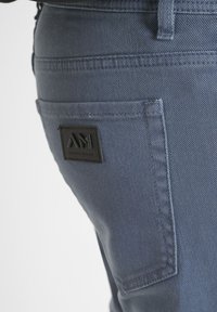 Primer plano de un bolsillo trasero de unos pantalones vaqueros azules con un parche rectangular negro de logo de Antony Morato cosido.
