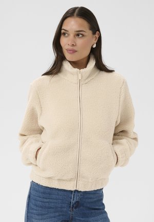 Femme portant une veste polaire beige à fermeture éclair avec les mains dans les poches, associée à un jean bleu, regardant légèrement sur le côté.