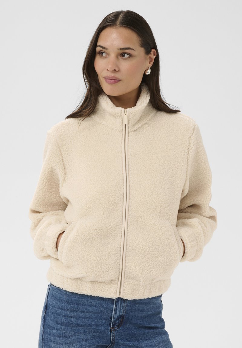 Femme portant une veste polaire beige à fermeture éclair avec les mains dans les poches, associée à un jean bleu, regardant légèrement sur le côté.