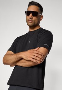Black short-sleeve T-shirt with a chest pocket and white logo on the sleeve. Model wears oversized black sunglasses, arms crossed.