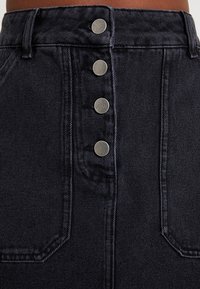 Jupe en denim noire taille haute avec cinq boutons-pression en argent, poches diagonales et un tissu texturé aux coutures visibles.