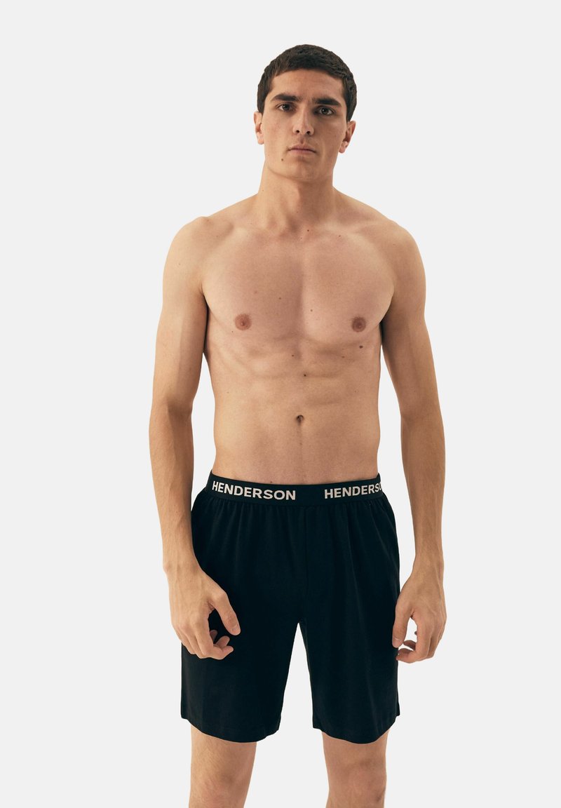 Czarne bokserki z elastycznym pasem, na którym widnieje napis "HENDERSON" w białym kolorze. Model stoi z neutralnym wyrazem twarzy, eksponując wysportowaną sylwetkę.