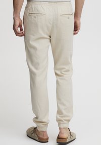 Pantalones de lino beige claro con cintura ajustable, puños elásticos y dos bolsillos traseros, con un acabado de tela suave y texturizada.