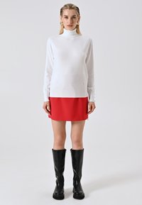 Frau in einem weißen Rollkragenpullover, rotem Minirock, großen goldenen Ohrringen und schwarzen Stiefeln bis zum Knie, die vor einem schlichten Hintergrund steht.