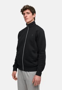 Svart zippered hettegenser med høy krage, laget av mykt stoff, med en subtil logo på brystet og elastiske mansjetter. Brukt med grå bukser.