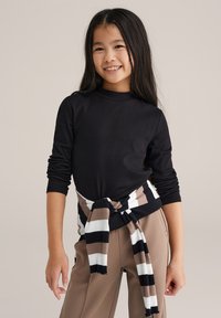 Fekete hosszú ujjú felső, bordázott textúrával. A derék körül barna-fehér csíkos pulóver megkötve. Barna, sima felületű nadrág.