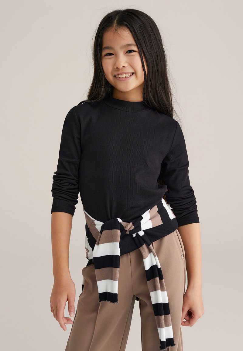 Fekete hosszú ujjú felső, bordázott textúrával. A derék körül barna-fehér csíkos pulóver megkötve. Barna, sima felületű nadrág.