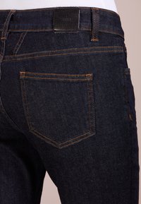 Tmavě modré džíny s úhlovými zadními kapsami s oranžovým prošíváním a černou koženou nášivkou připevněnou na pasu.