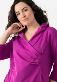 Magenta-Top mit Drapierung am Kragen, langem Ärmel und Faltenwurf am Ausschnitt. Glatte Stofftextur, taillierte Silhouette.