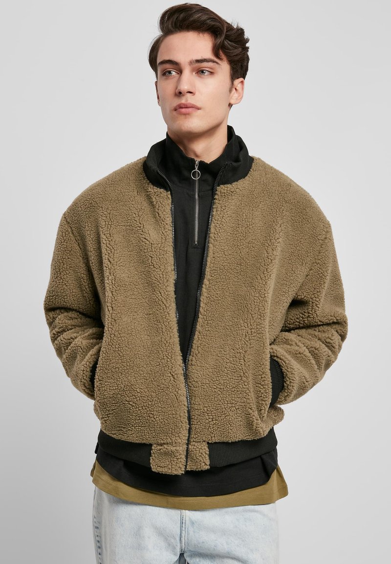 Bombažna jakna iz umetnega ovčjega kožuha v olivno zeleni barvi z visokim črnim ovratnikom, zadrgo in rebrastimi manšetami, nošena preko slojevite črne in olivno zelene majice.