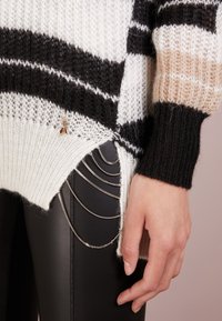 Maglione a righe nei colori nero, bianco e beige. Presenta uno spacco laterale e catene decorative sui pantaloni neri. La texture è morbida e leggera.
