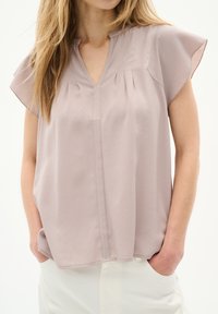 Blusa a maniche corte in morbido tessuto mauve, con scollatura a V, pieghe e vestibilità rilassata, abbinata a pantaloni chiari.