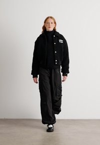 Veste en polaire noire courte avec une finition texturée, dotée de boutons argentés et d'un logo. Associée à un pantalon cargo noir ample et des chaussures noires.