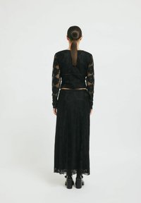 Schwarzes Spitzenoberteil mit langen Ärmeln und floralem Muster, kombiniert mit einem passenden langen Rock. Beide Teile verfügen über eine weiche Textur und eine taillierte Passform. Sichtbare schwarze Stiefeletten mit Absätzen.