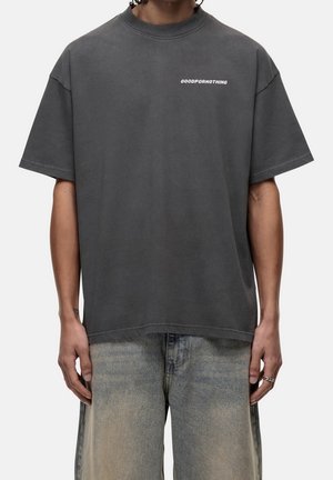Osoba nosząca luźny ciemnoszary t-shirt z napisem „GOODFORNOTHING” na piersi, w połączeniu z wyblakłymi jasnoniebieskimi jeansami.