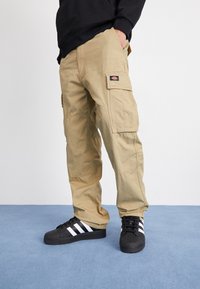 Pantaloni cargo beige realizzati in tessuto resistente, caratterizzati da molteplici tasche, cuciture rinforzate e un logo a toppa sulla coscia. Indossati con sneakers nere.